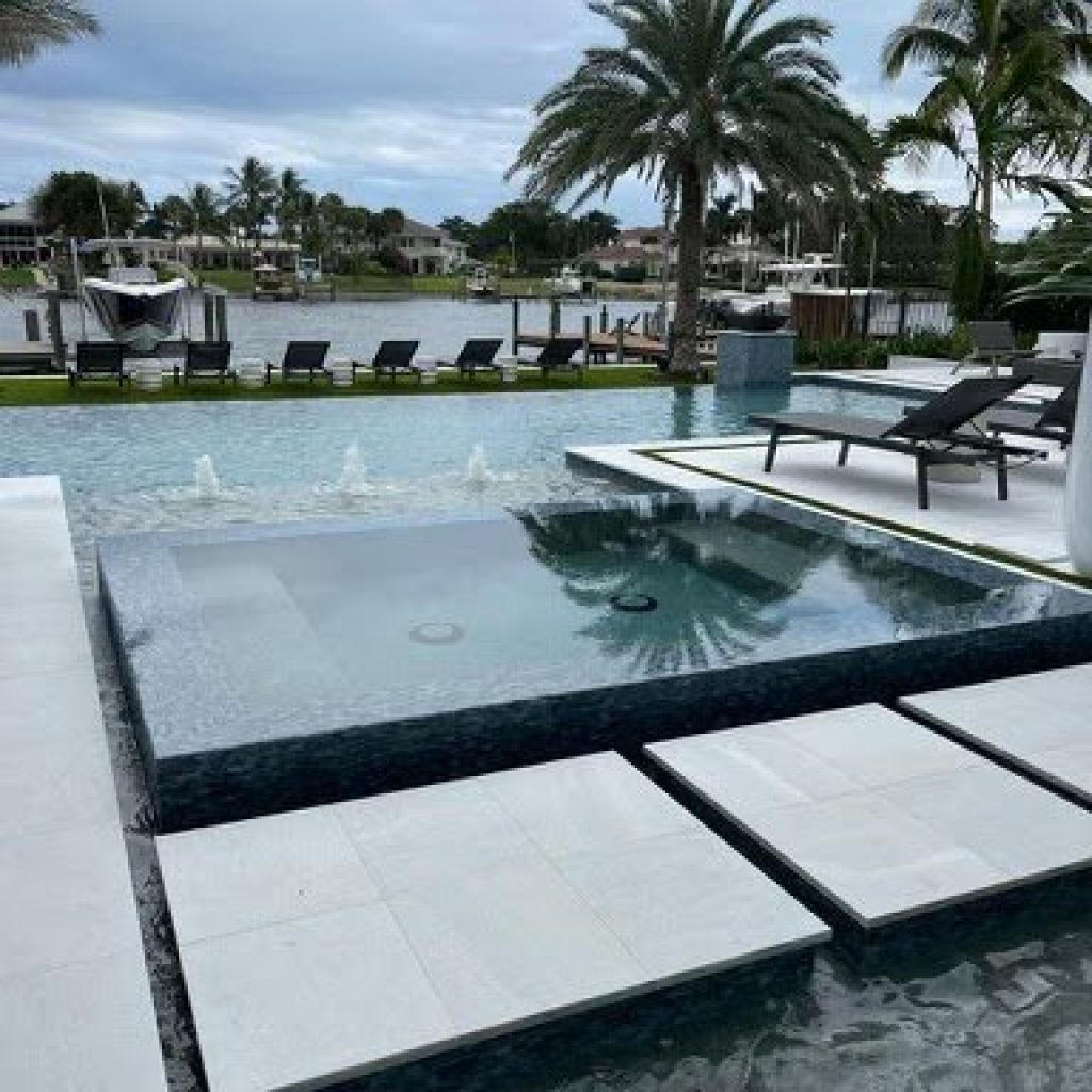 Infinity-edge spa spilling into a waterfront pool with stepping pads and lounge chairs