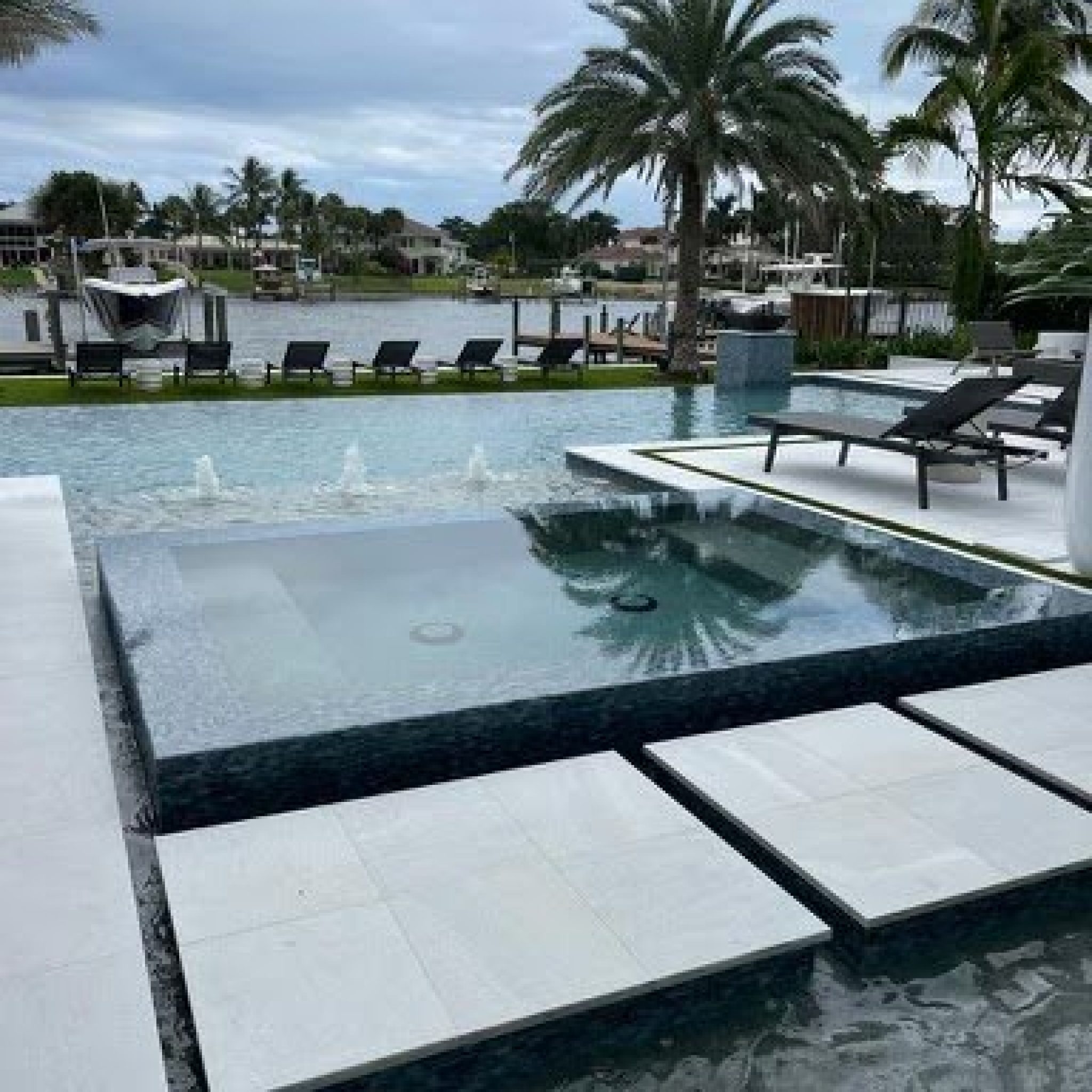 Infinity-edge spa spilling into a waterfront pool with stepping pads and lounge chairs