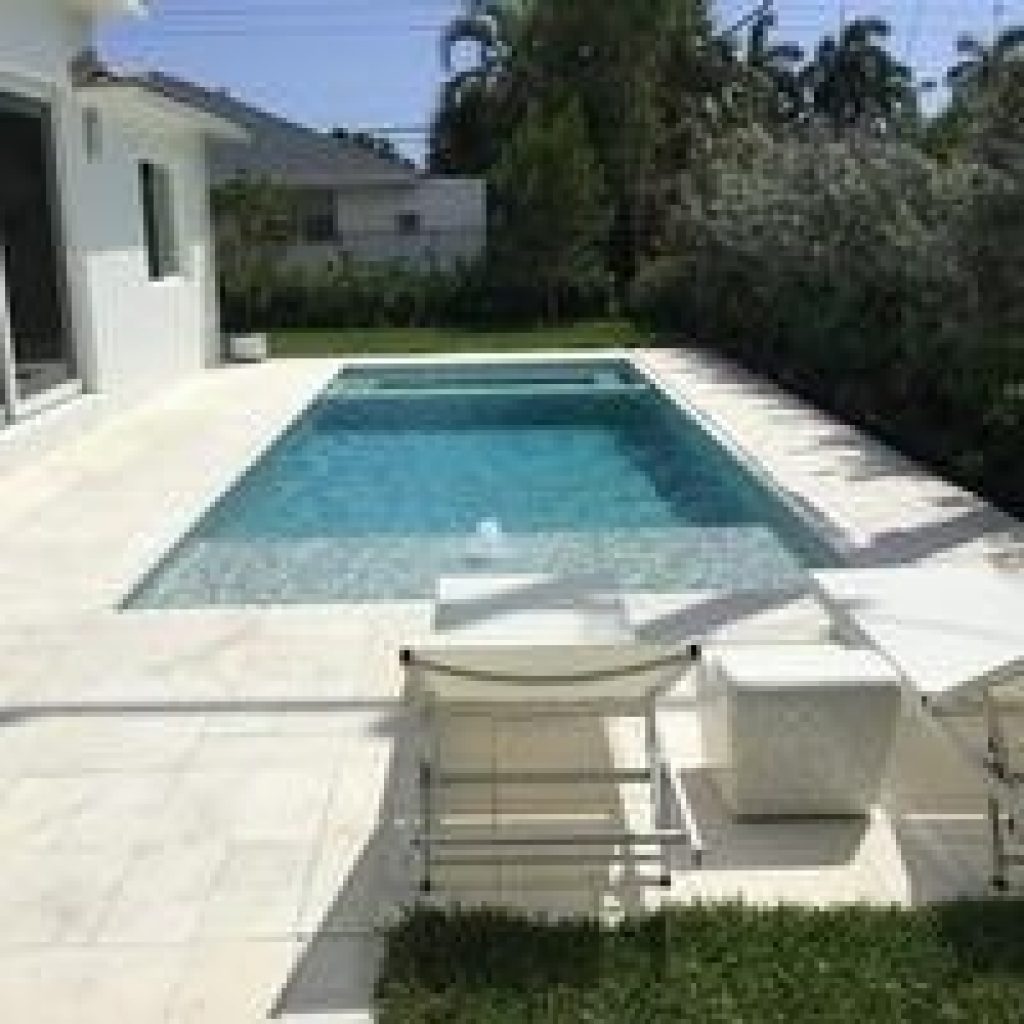 Modern rectangular pool with a bubbler sun shelf and white lounge chairs beside a white home