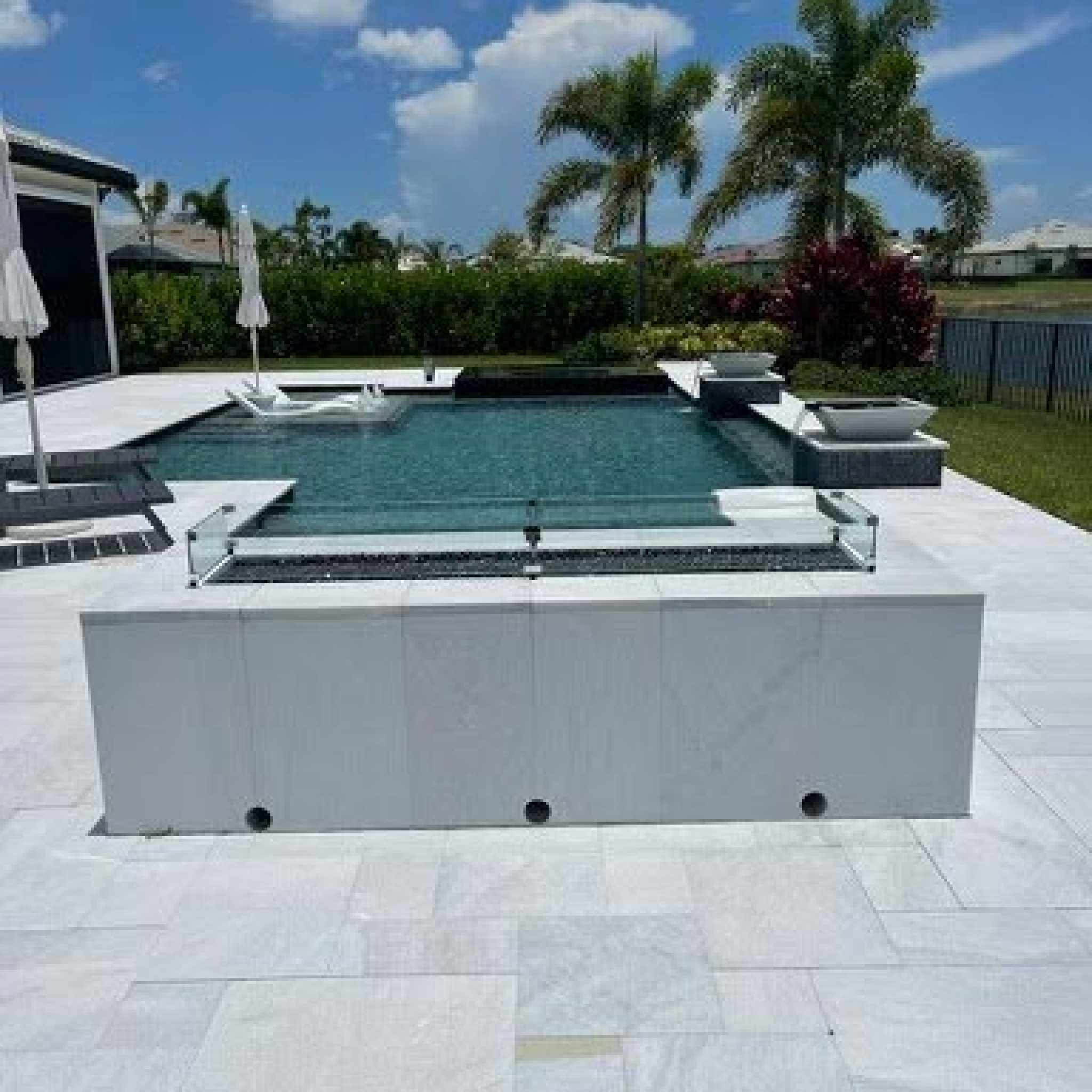 Raised spa with a white porcelain wall and glass fire feature beside a pool with sun shelf loungers