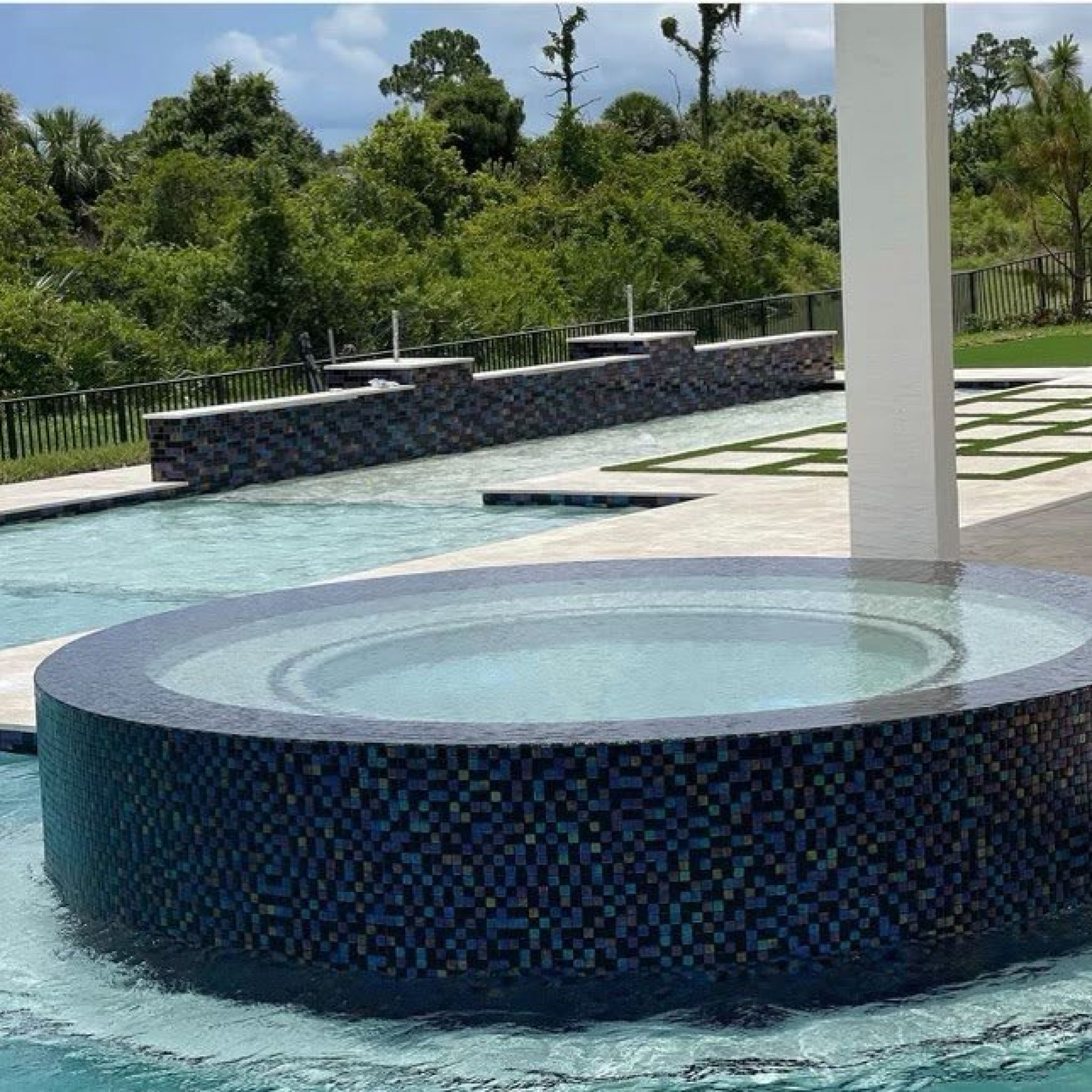Round raised spa finished in dark glass mosaic tile beside a pool