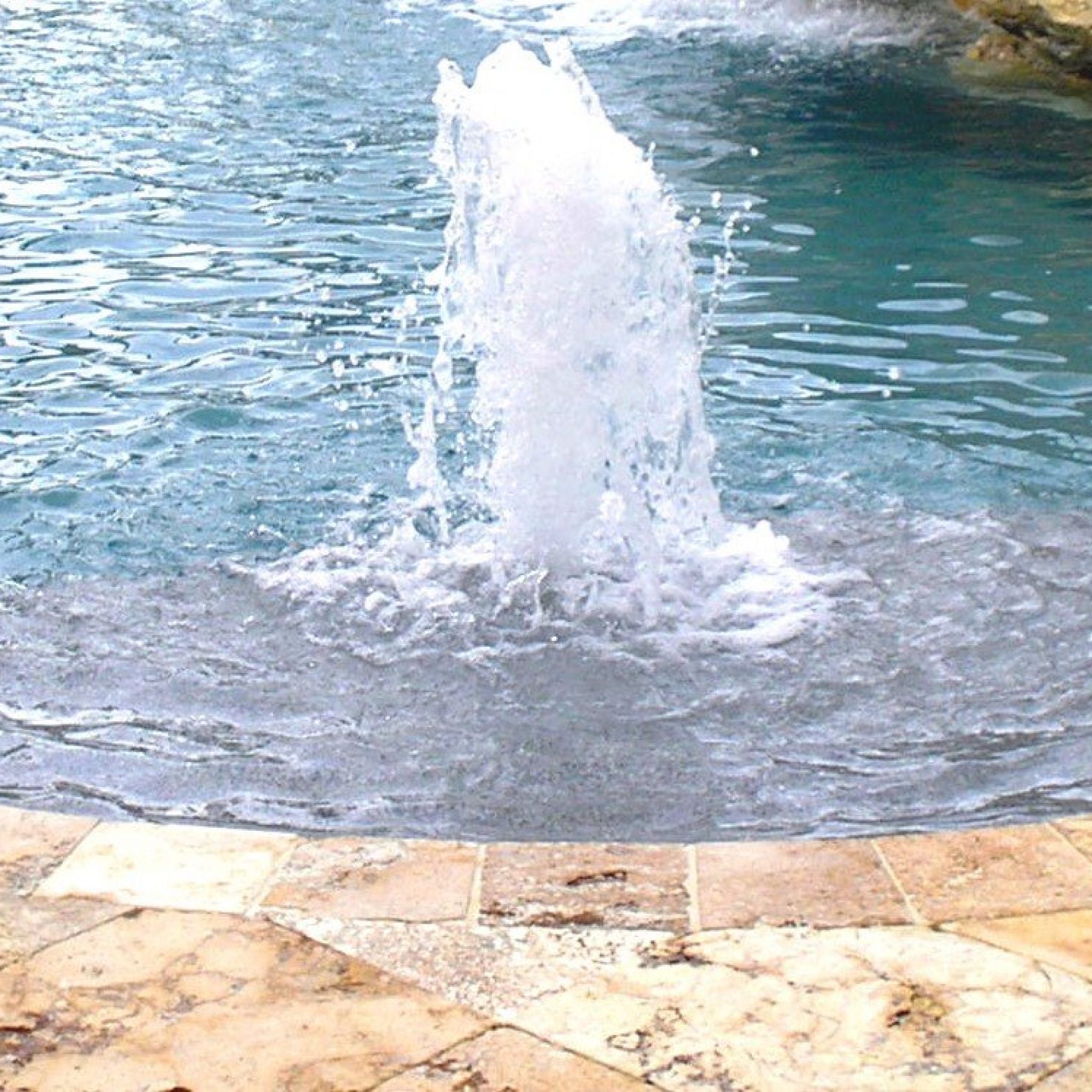 Bubbler fountain spraying water on a pool sun shelf with travertine coping